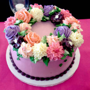 Photo Apr 05, 3 13 25 PM A round cake with purple frosting, decorated with pink, white, and purple buttercream flowers and green leaves, sits on a white base. A glass sits behind the cake on a pink and black tablecloth.