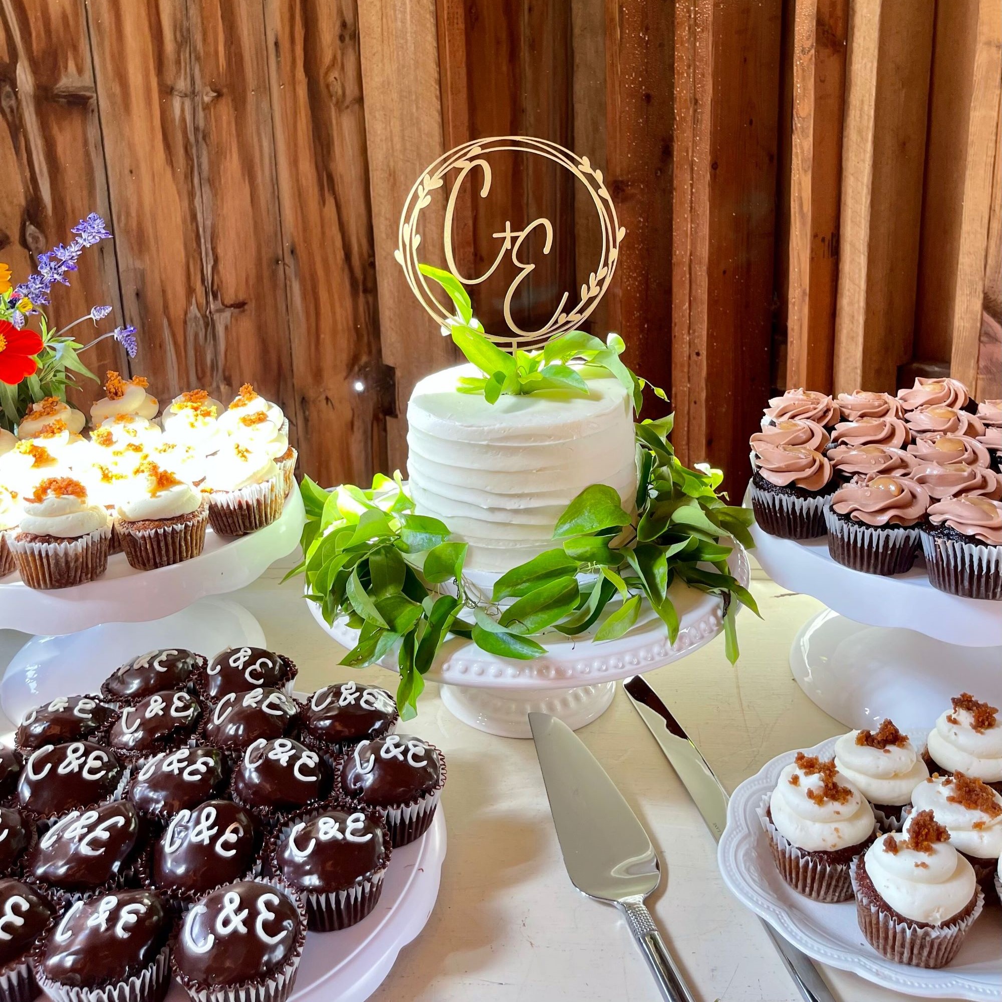 A white cake with a C & E topper and green leaves is surrounded by assorted cupcakes on stands, displayed on a table with a cake server and knife, set against wooden walls.