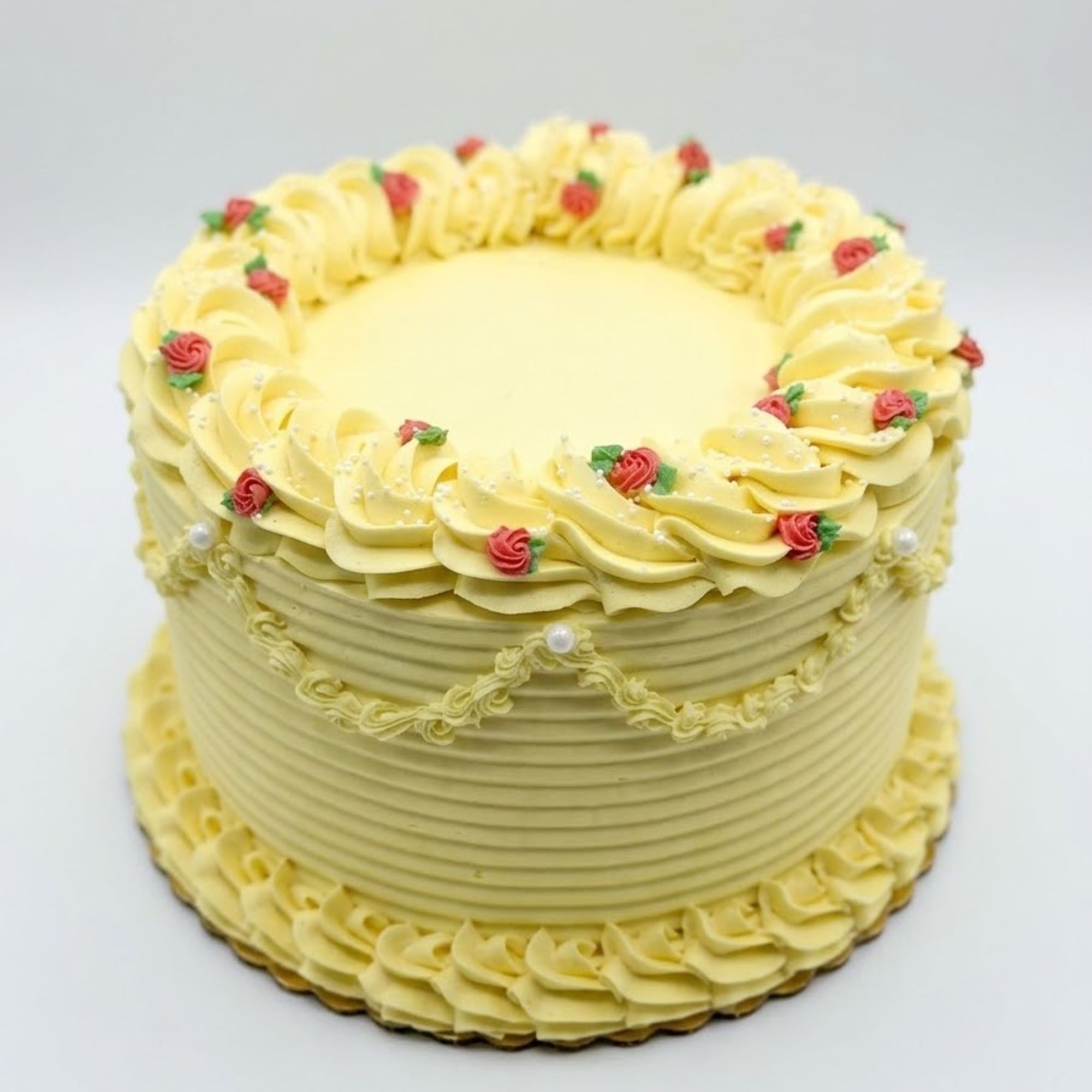 A round yellow cake with piped buttercream swirls, small pink and green flower decorations, and pearl accents, set against a plain white background.