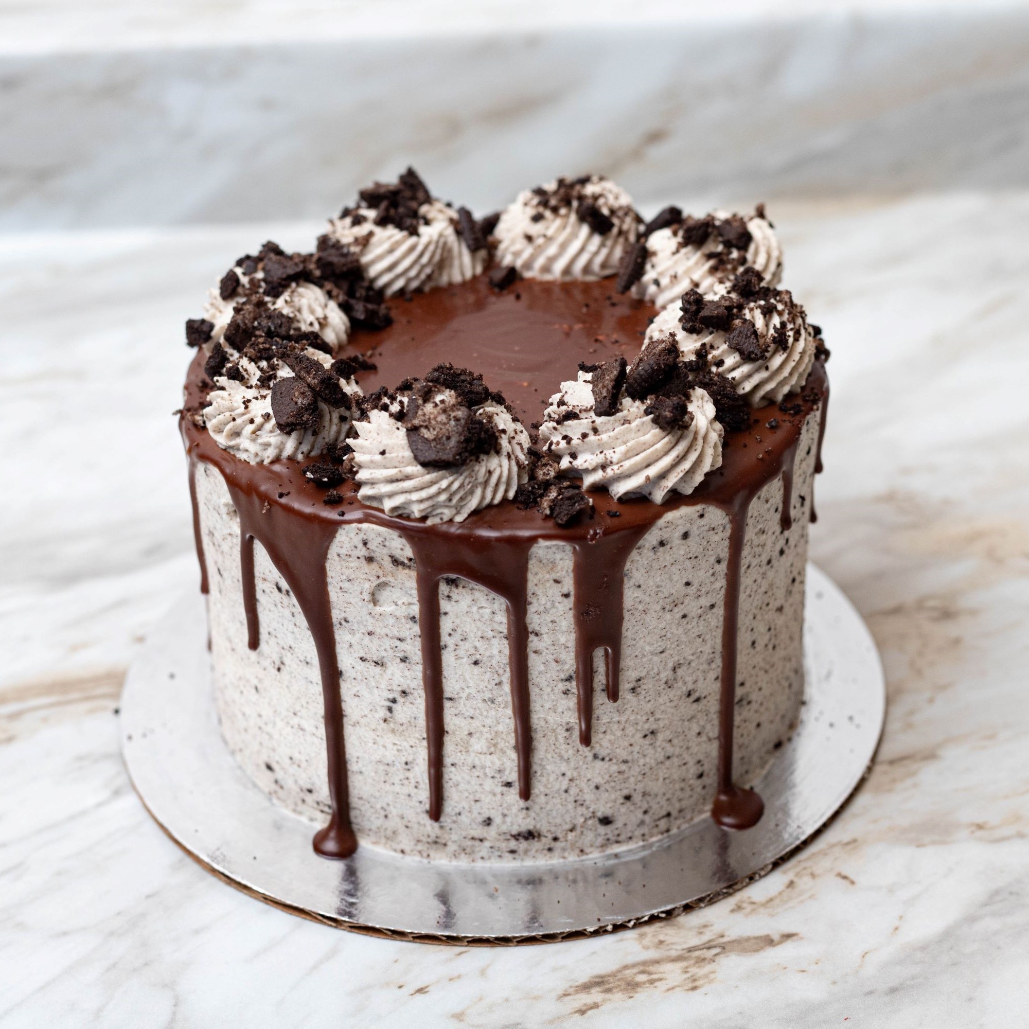 A cookies and cream cake with chocolate ganache dripping down the sides, topped with whipped cream swirls and crushed chocolate cookies on a marble surface.
