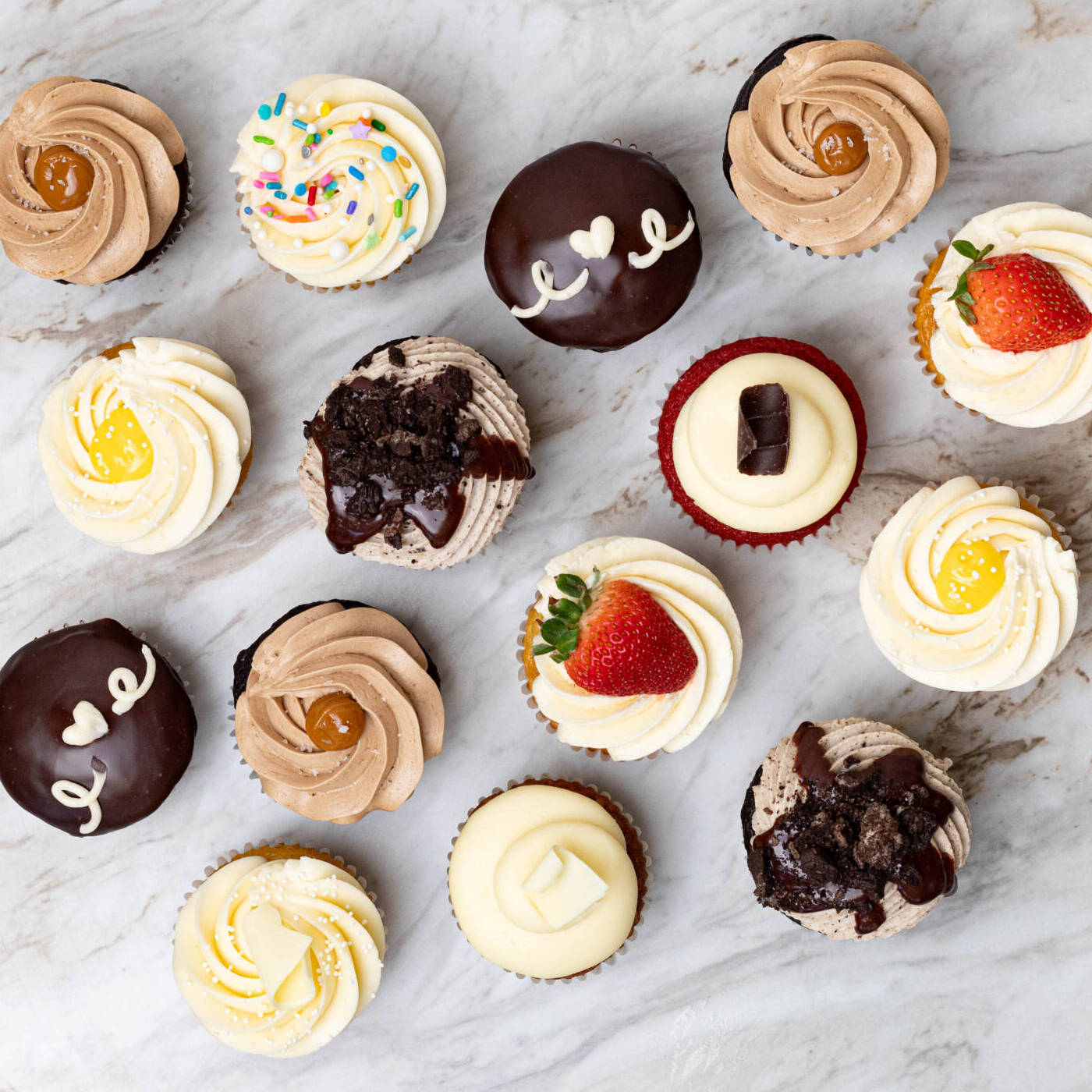 A variety of twelve cupcakes with different frostings, toppings, and fruit arranged on a marble surface.